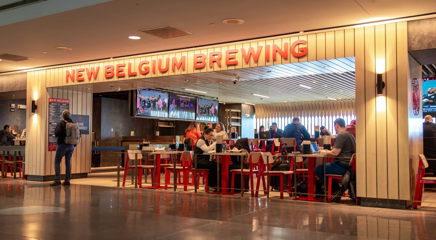 Pintu masuk ke New Belgium Brewing di Bandara Internasional Denver.  Ada bar olahraga dengan TV di sebelah kiri dan banyak meja dan kursi merah dengan orang-orang yang makan di sebelah kanan