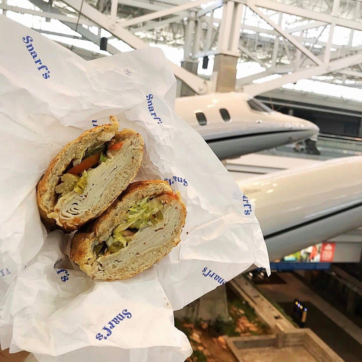 Snarf's Sandwich cut in half in white paper, held up in front of airplane in Denver International Airport