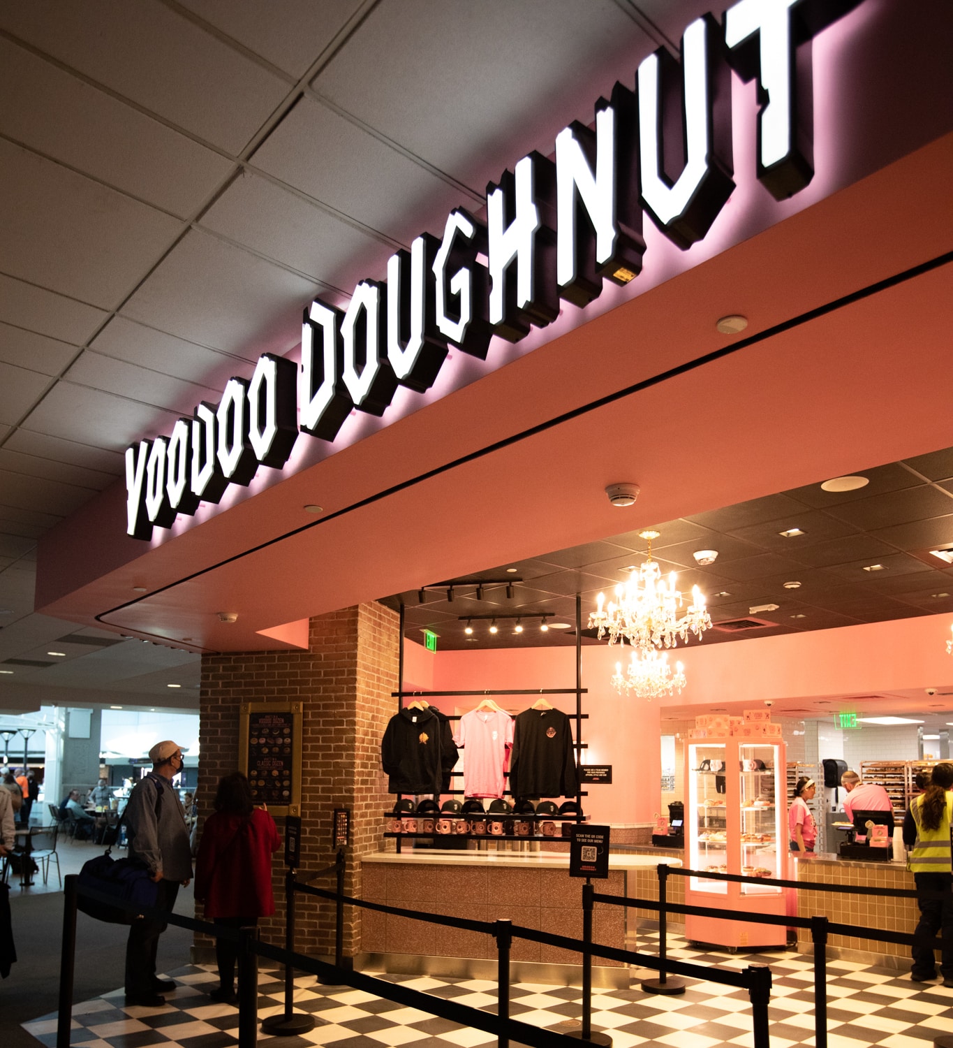 Etalase Voodoo Donut di Bandara Internasional Denver.  Dinding bercat pink dengan lantai kotak-kotak hitam putih