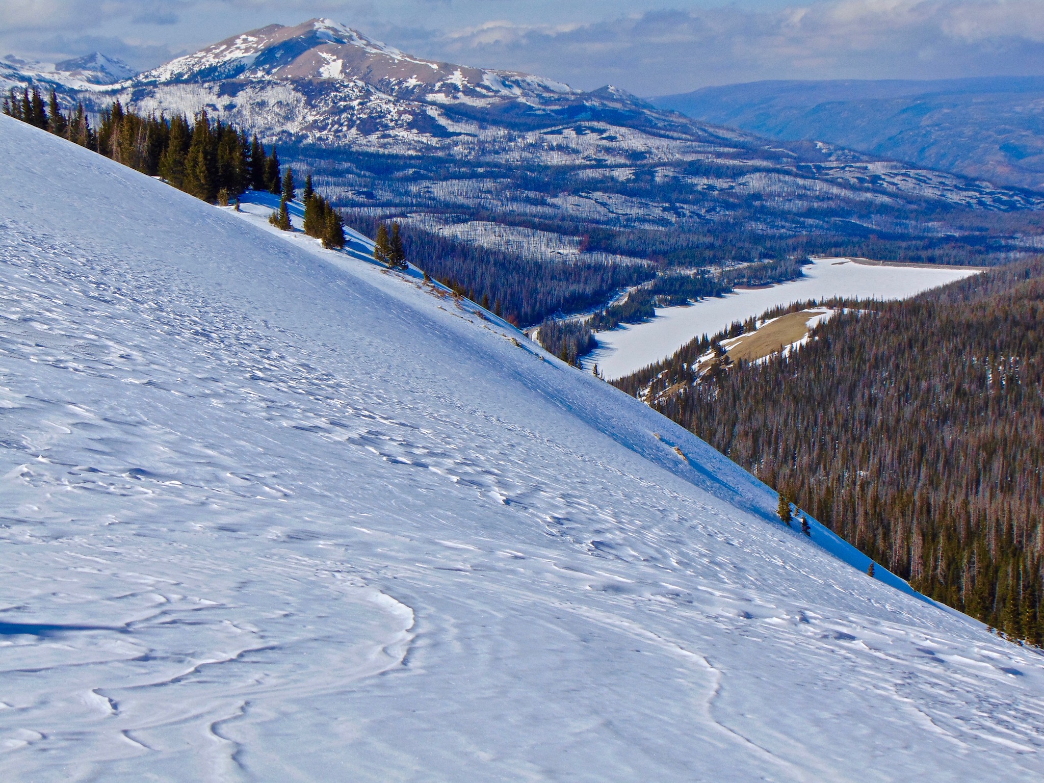 A steep snow slope with long distance views of Cameron Peak with its bald summit and forests around its lower slopes.