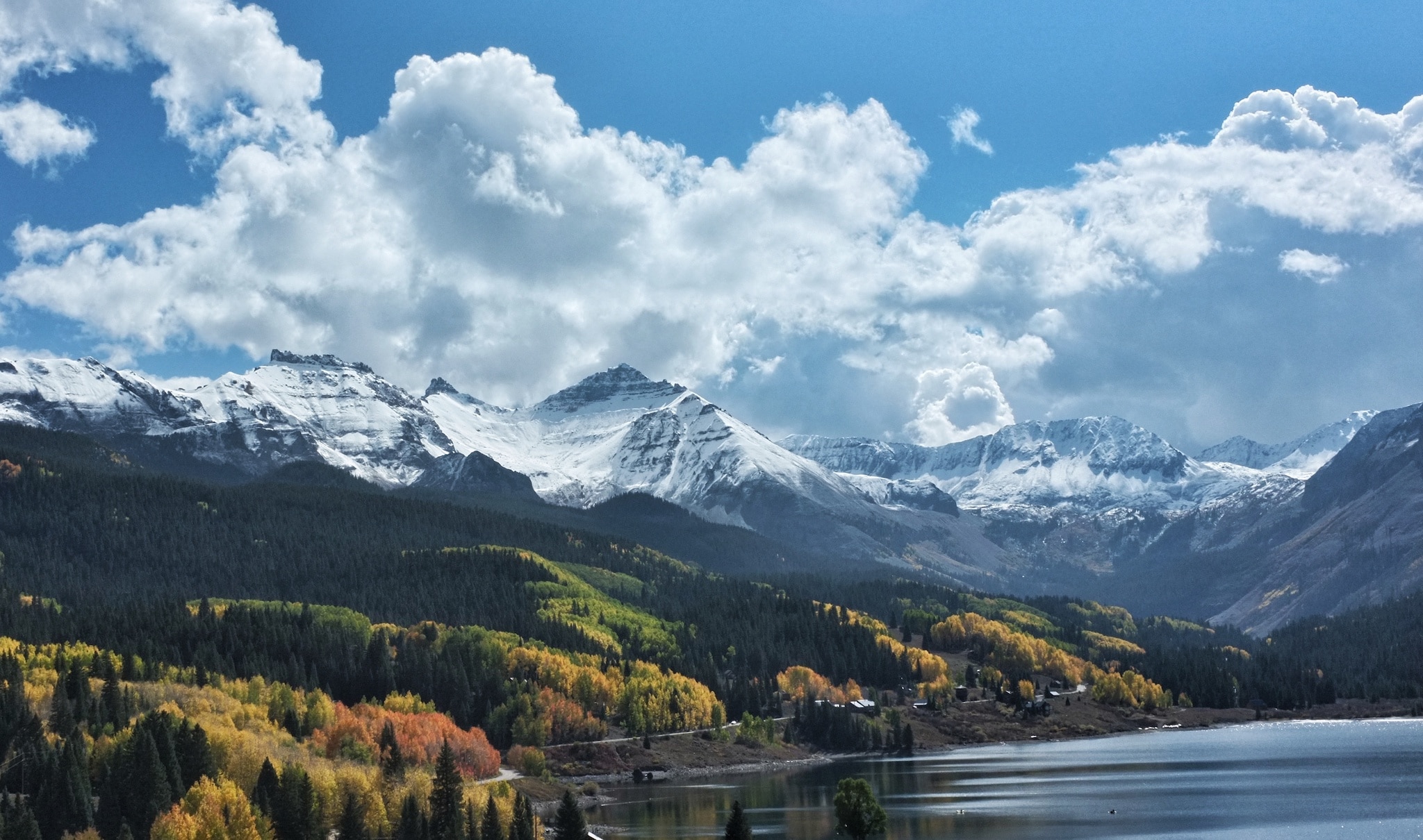 A clear blue lake surrounded by towering snow capped mountains and autumn leaves.