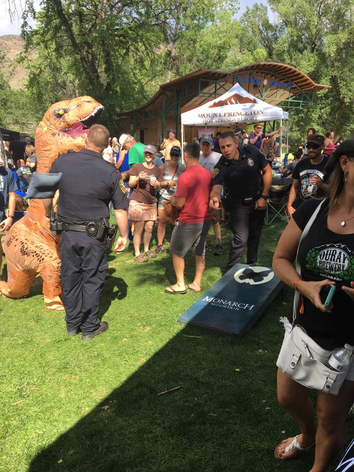 Police offer throwing a bean bag onto a cornhole board in the middle of a crowd of people gathered around on the grass. Everyone is attenting the annual Colorado Brewers Rendevous in downtown Salida