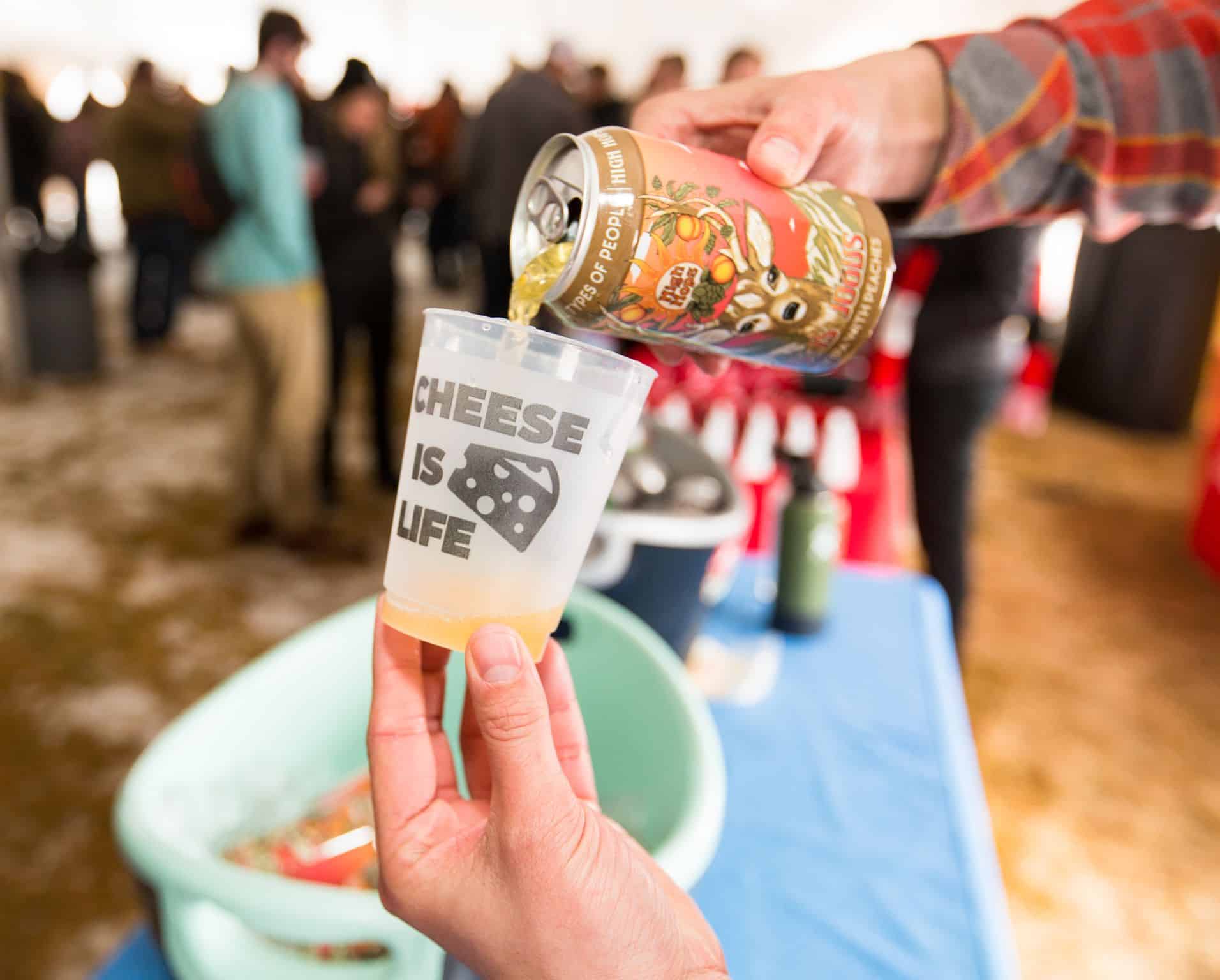 Pouring a can of craft ber into a cup that says Cheese is Life at the Mile High Grilled Cheese and Mac festival in Denver