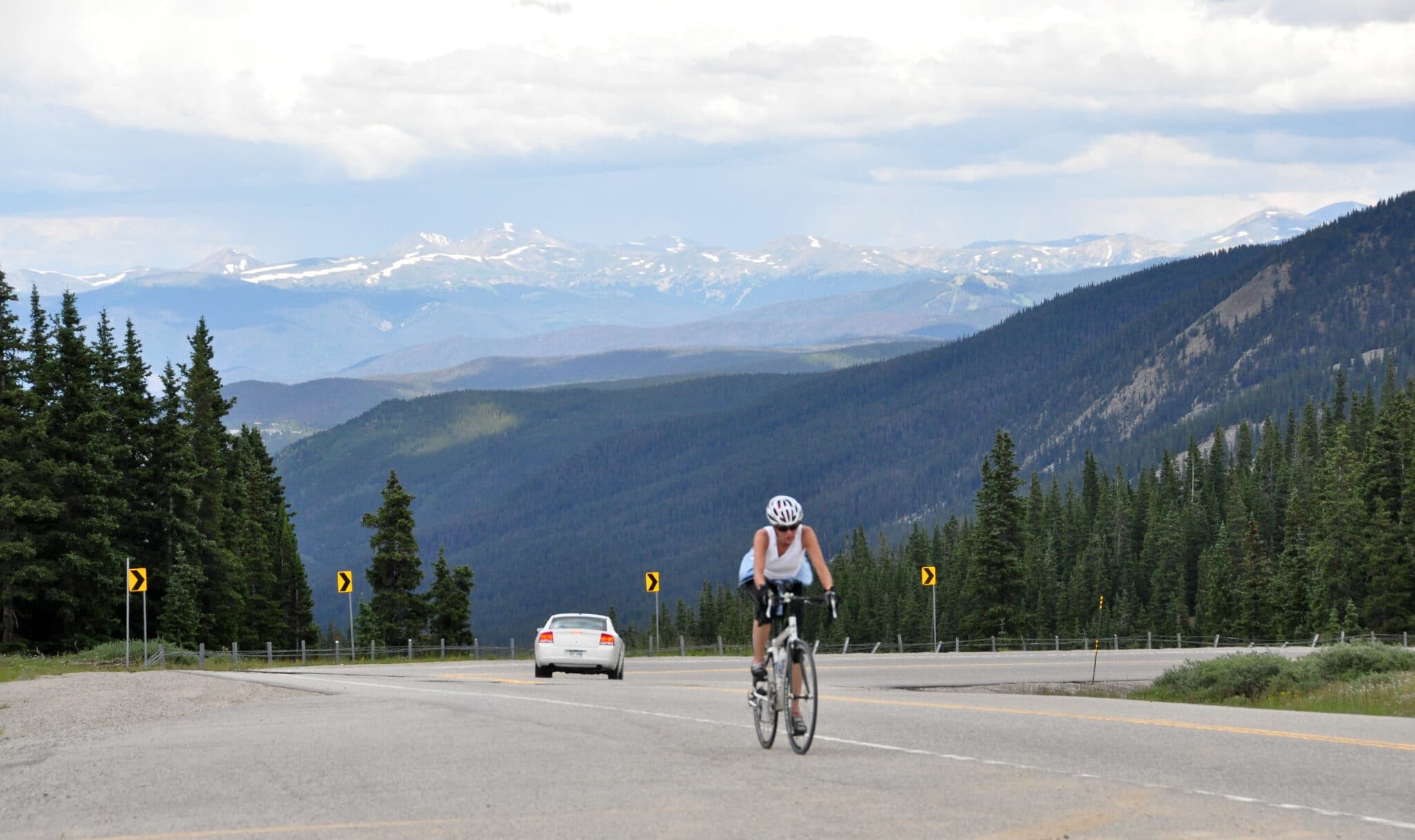 A paved mountain pass with a cyclist climbing up and a car descending, a large valley and distant mountains with patchy snow are visible.