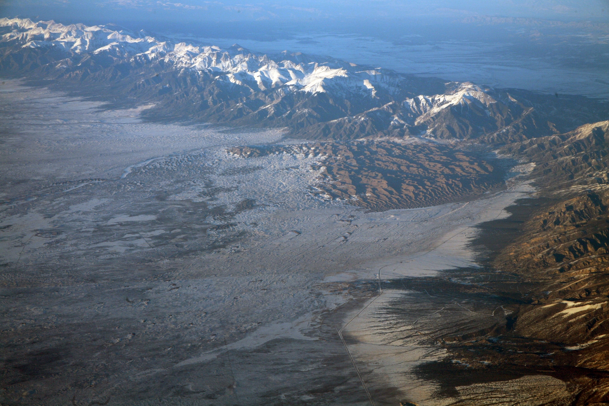 Aerial photo looking down on a large, arid valley, a clump of sanddunes steeped in shades of brown, and a thin snowcapped mountain range behind it all.