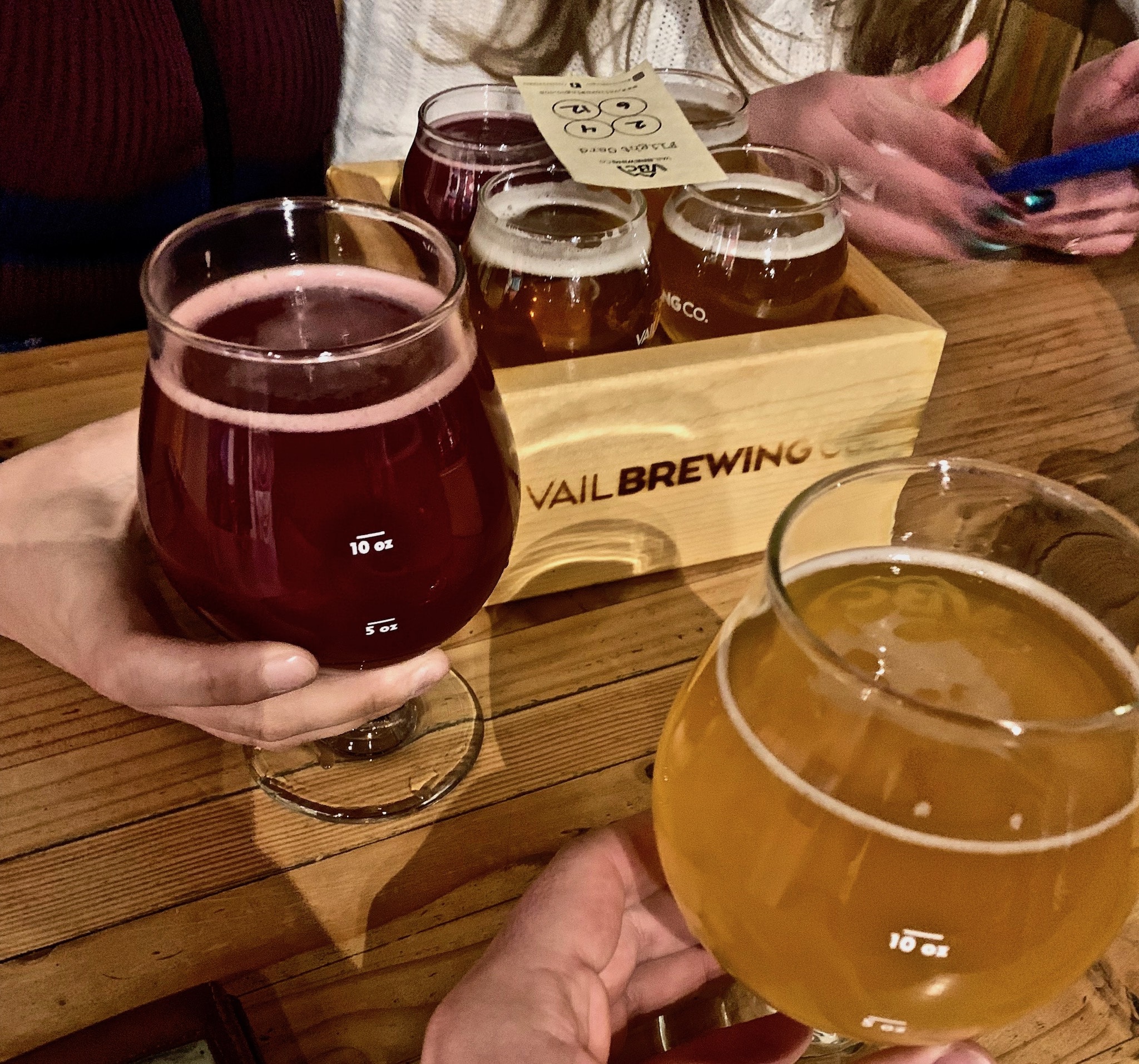 A table surface with two pints of beer in hands, and a small wooden box with 4, 2.5 ounce beer tasters.