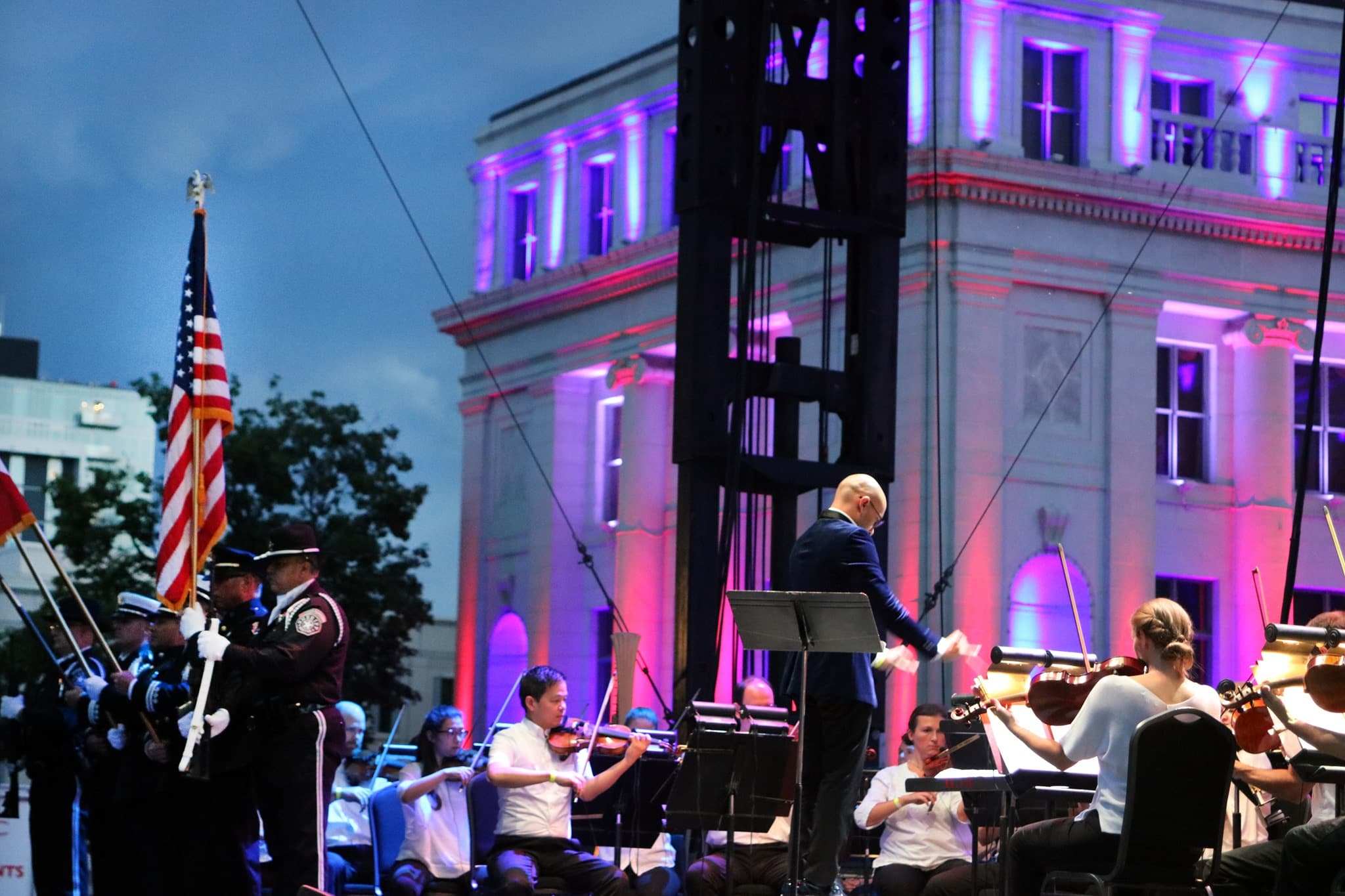 Conductor for the Colorado Symphony standing in front of his performers with a lit up white building in downtown Denver in the background. 