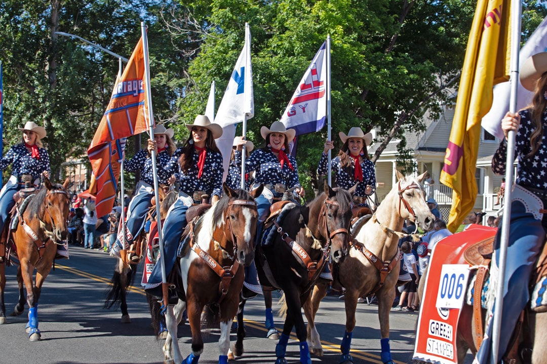 Independence Day parade in Greeley, Colorado. 3 people on horses holding flags in in the parade procession.