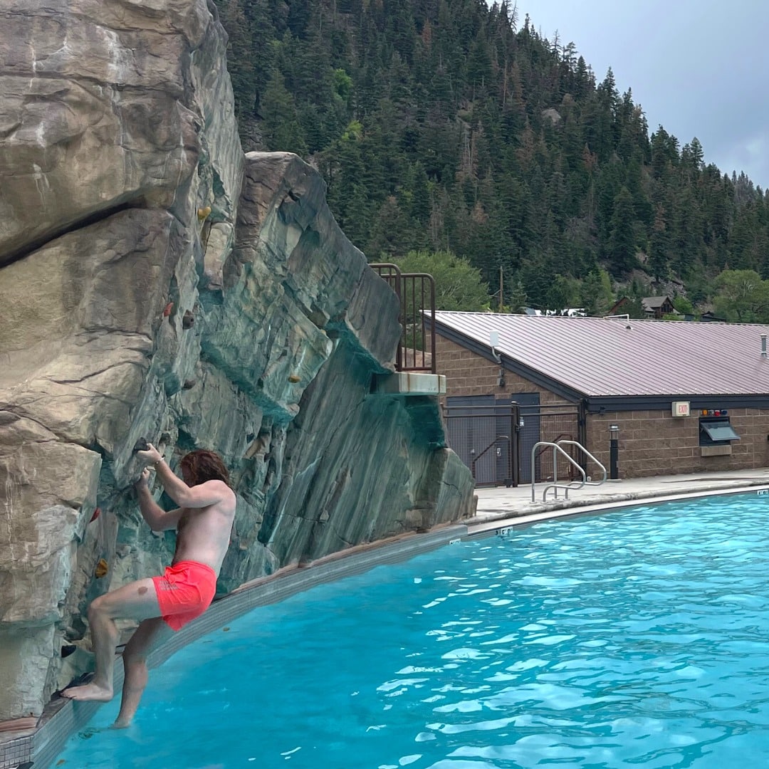 Person on an outdoor climbing wall over a pool.