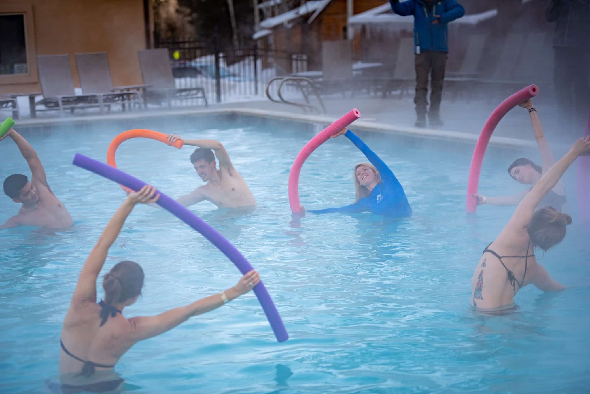 People holding pool noodles over their head while half submerged in a hot springs pool during a workout class. 