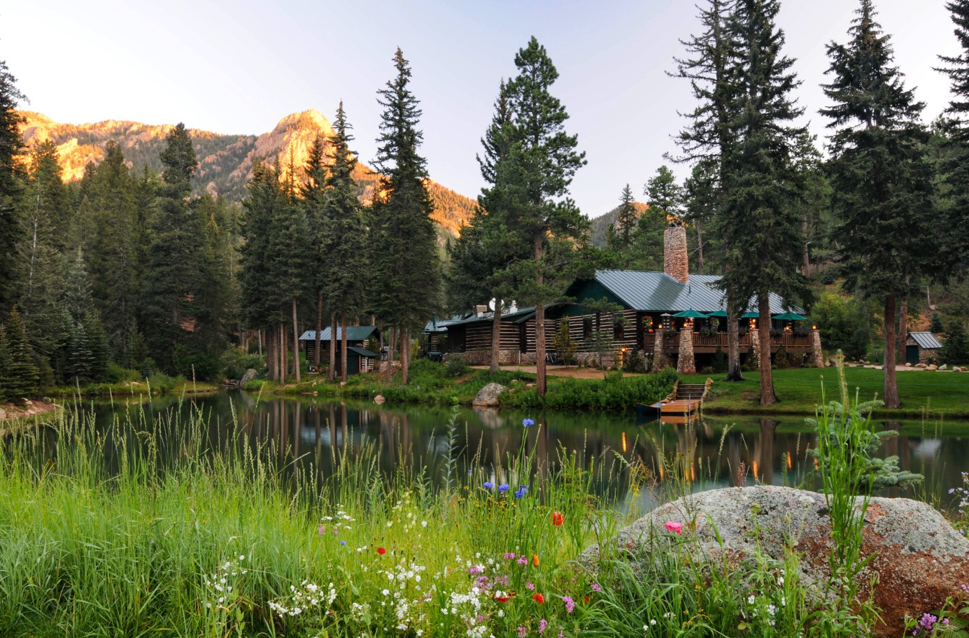 Log house sitting on the shore of a small pond surrounded by long green grass, evergreen trees, and mountains in the background at The Ranch in Emerald Valley, owned by The Broadmoor