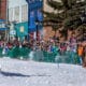 Man on horse pulls a man on skis over a jump built out of snow on the main street of a town