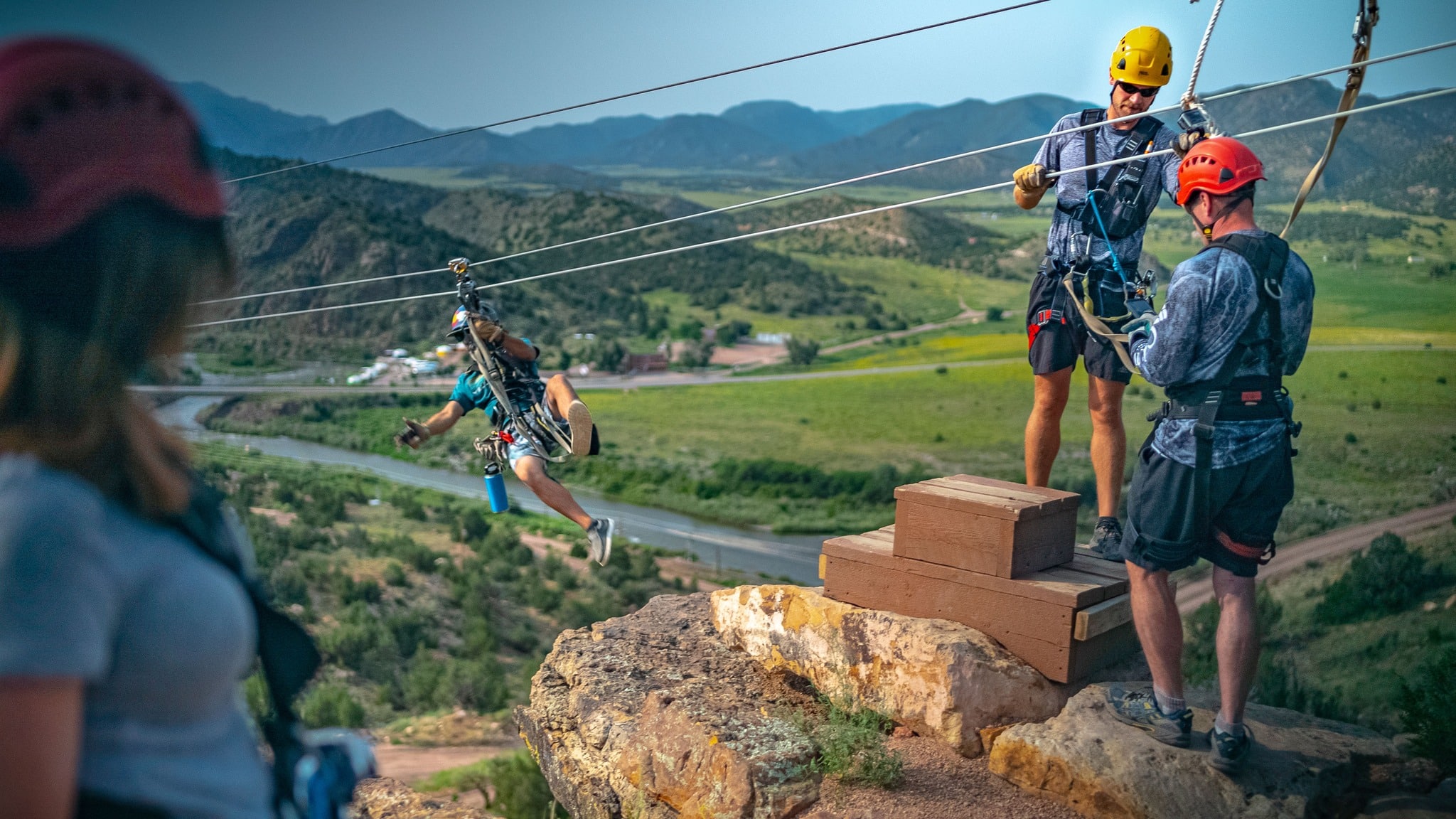 Three people at Royal Gorge Zipline Tours in red helmets standing on a wooden platform before connecting to the line to zip off