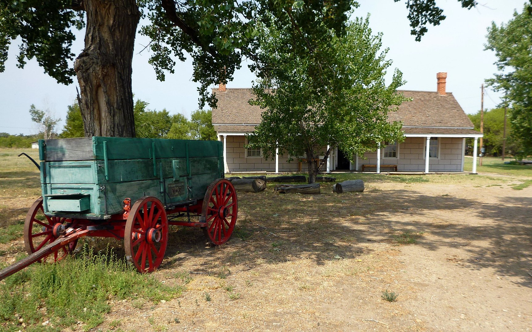 Old Boggsville house. One story home with a front porch and large tree with hanging branches in front of it. There is an old green wagon with red wheels in the foreground