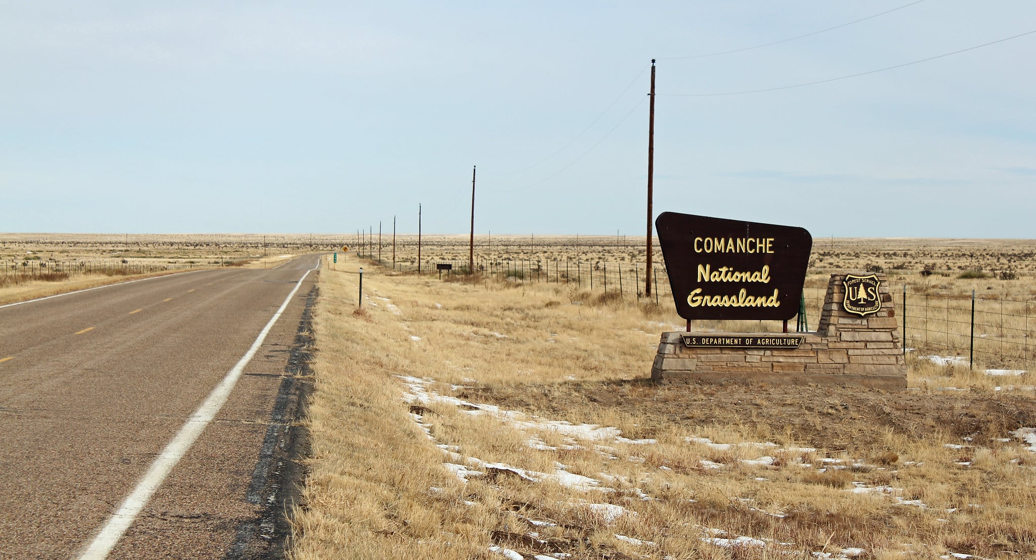 Long highway road with brown sign and yellowing lettering that says Comanche National Grasslands