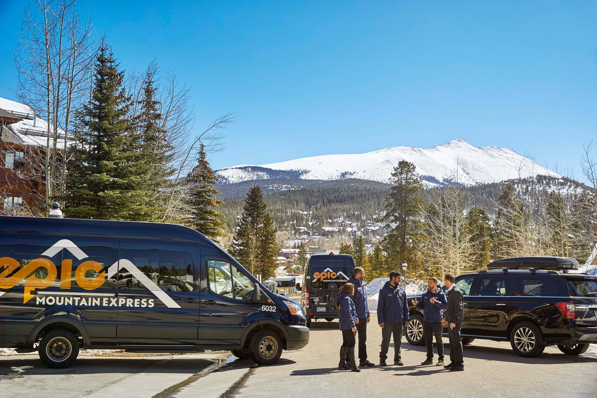 Three Epic Mountain Adventure black vans in a semi-circle and their drivers are talking. Snowy mountains and trees in the background