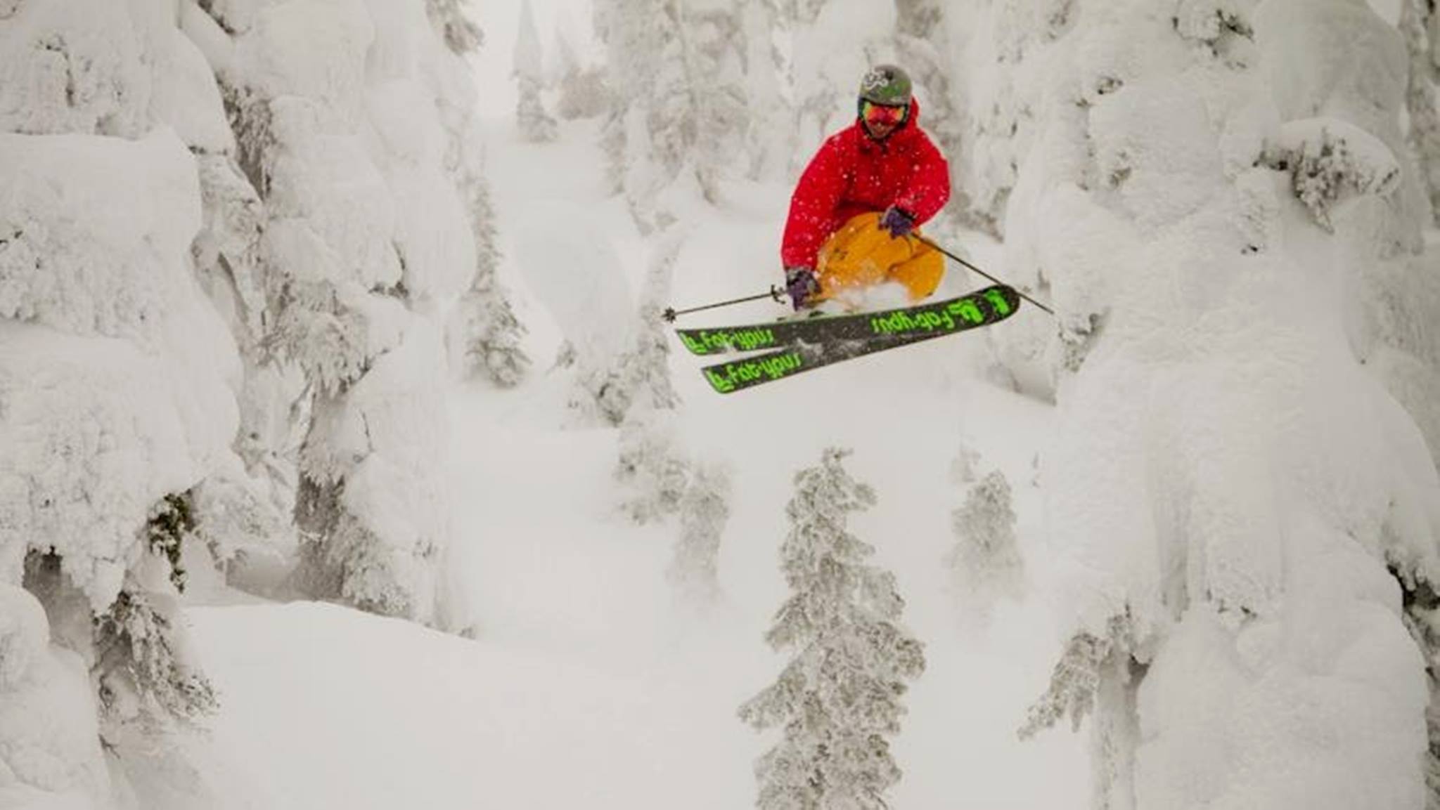 Skier in a red jacket catching air on black and green Fat-ypus Skis, made in Breckenridge. 