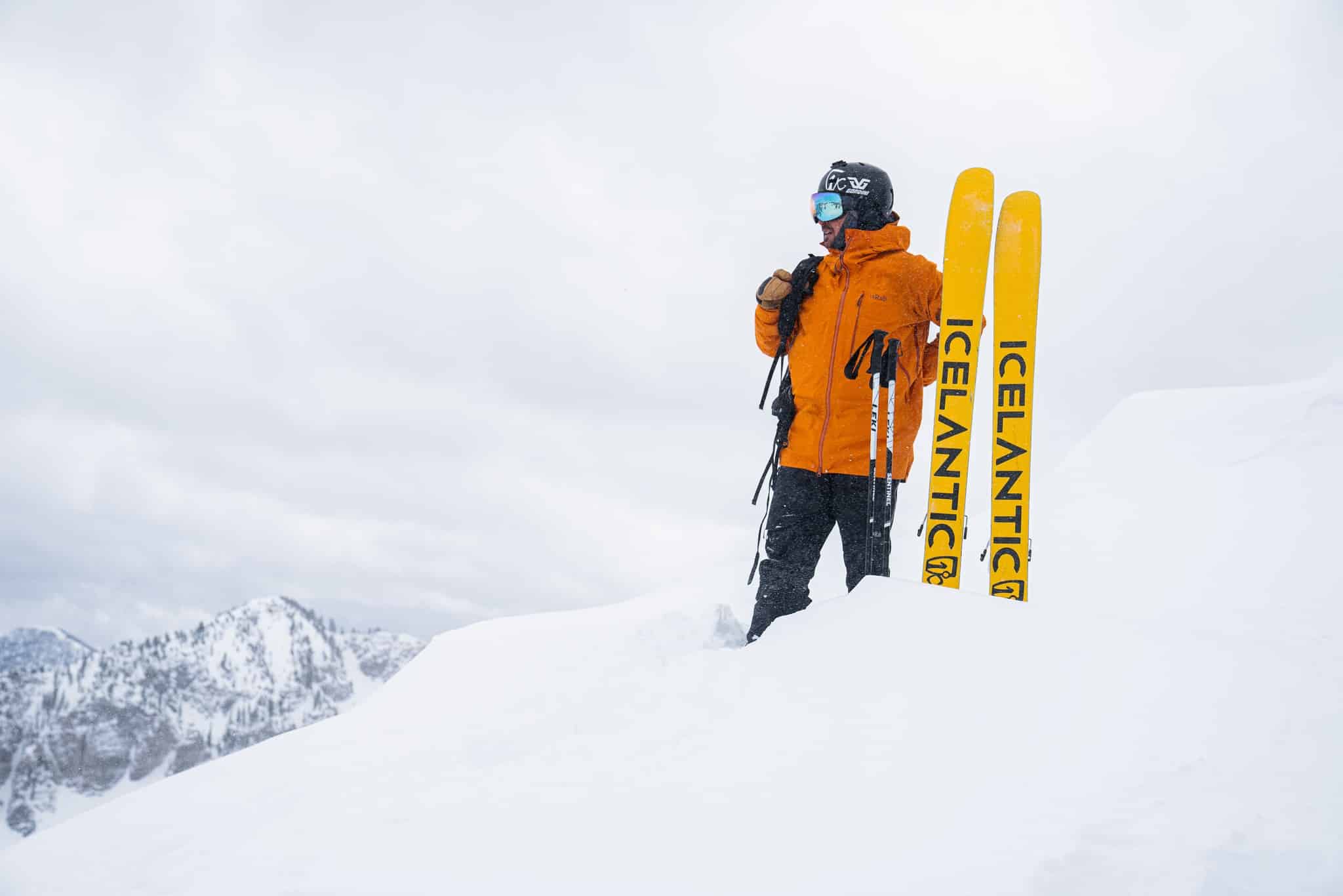 Pemain ski berjaket jingga cerah berdiri di puncak gunung bersalju di sebelah ski Islandia kuning