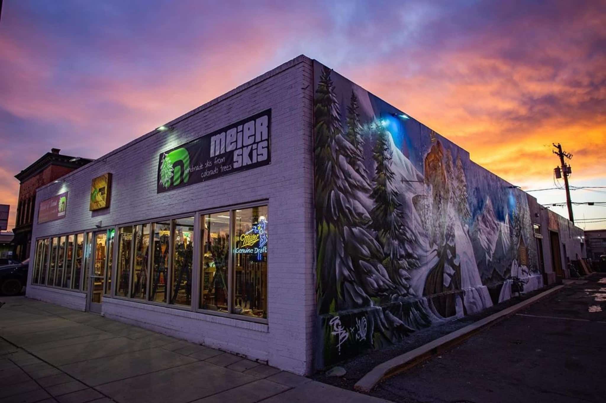 Matahari terbenam di atas gedung Meier Skis di pusat kota Denver.  Mural gunung besar di samping