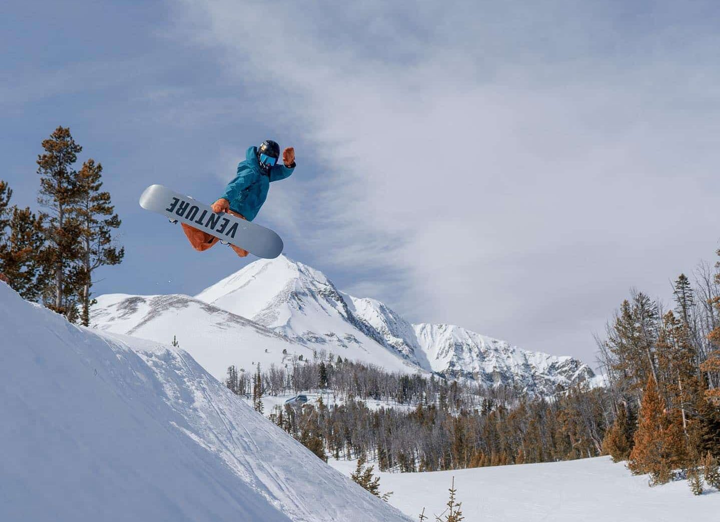 Snowboarder berjaket biru di udara setelah melompat di atas snowboard Venture putih