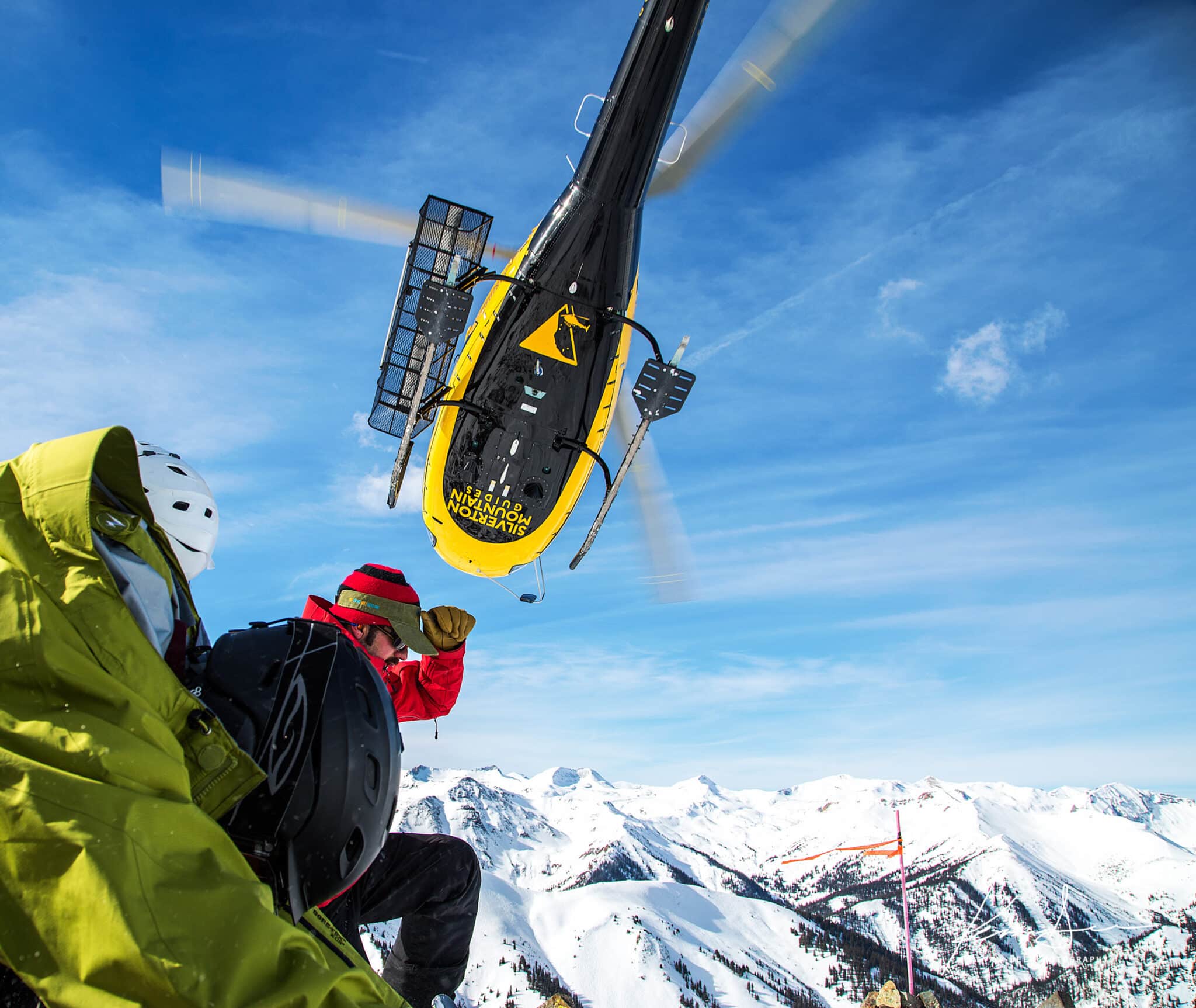 Yellow and black Silverton Mountain Guides helicopter hovers over 2 skiiers it dropped on the top of a mountain