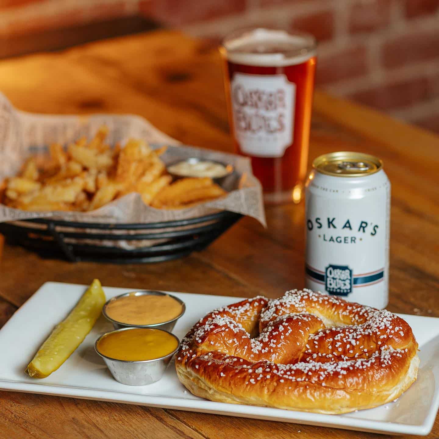 White platter with fluffy pretzel, cheese dip, and pickle spead in front of a basket of french fries with 2 Oskar Blues beers inbetween