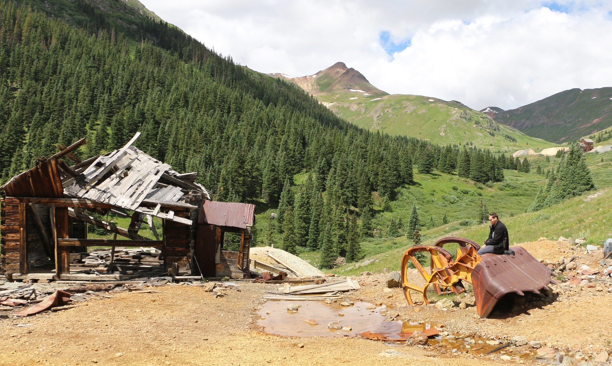 Old wooden building falling inward and man sitting on a rusted piece of farm equipment in the ghost town of Animas Forks