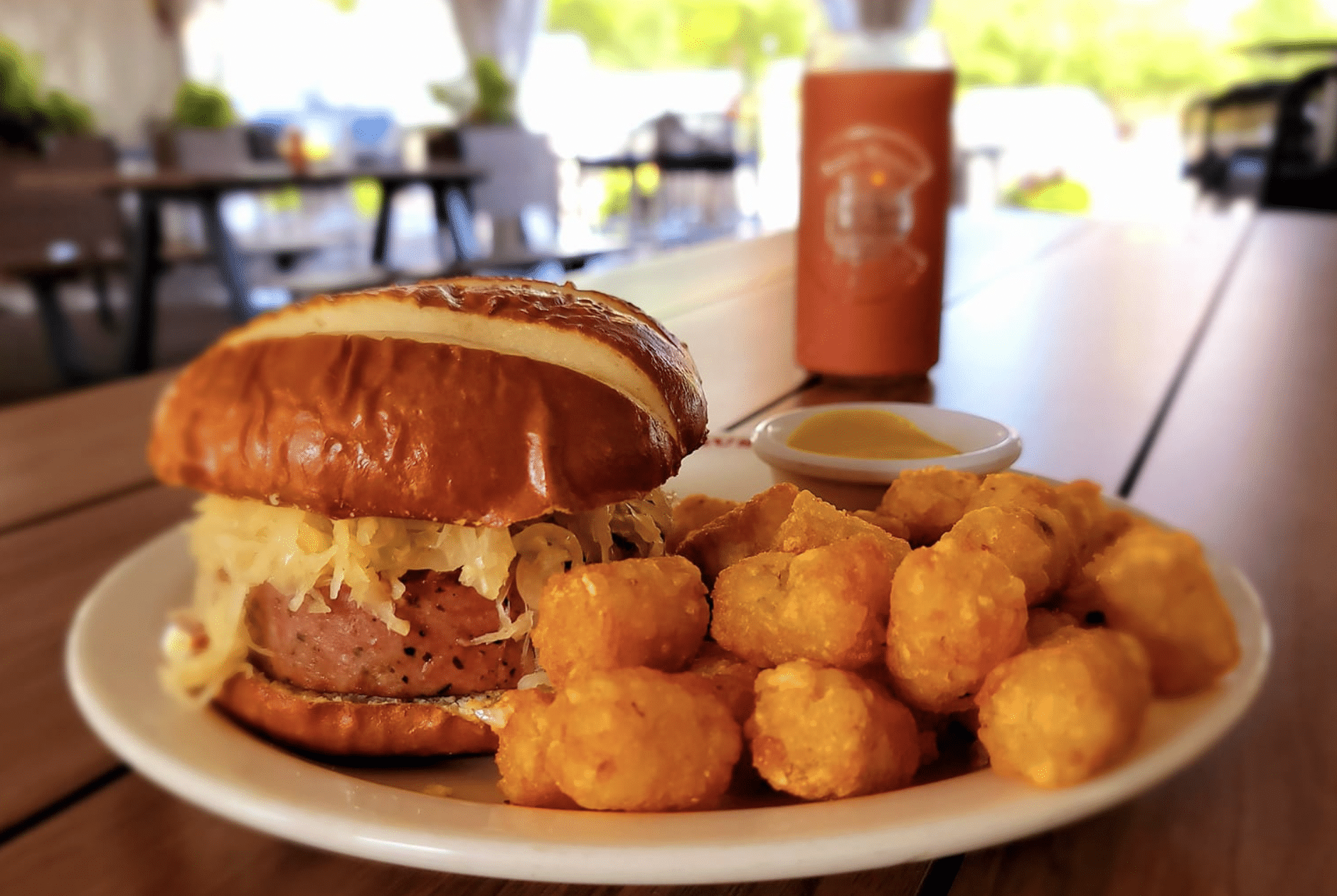 White plate with pretzel bun burger and tater tots with glass of beer in the background at Bull & Bush Brewery in Denver