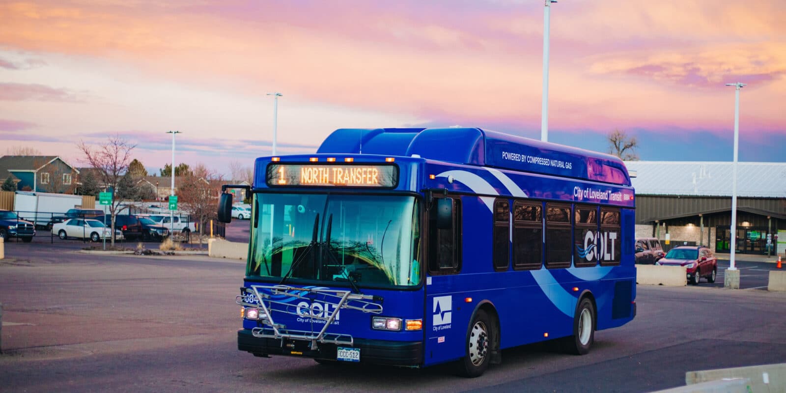 Blue City of Loveland Transit (COLT) bus driving on a road. Sky is orange and purple with a sunset