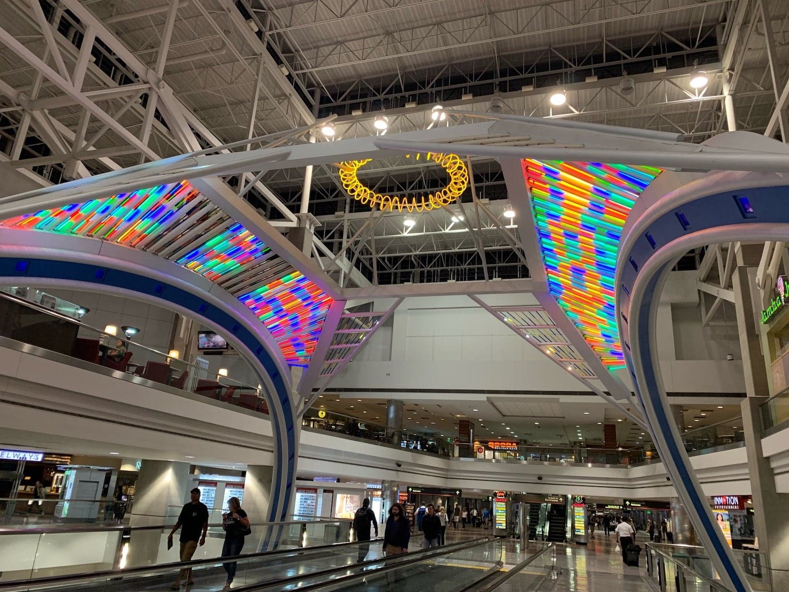 Pertokoan dan restoran Bandara Internasional Denver mengelilingi patung seni berwarna-warni di pusat Concourse B