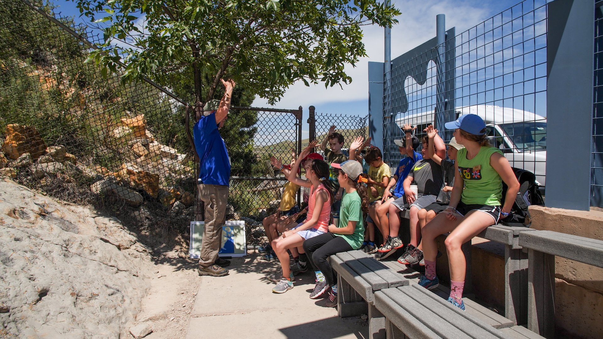 Group of children sitting on small set of bleachers while Dinosaur Ridge tour guide in blue polo explains something