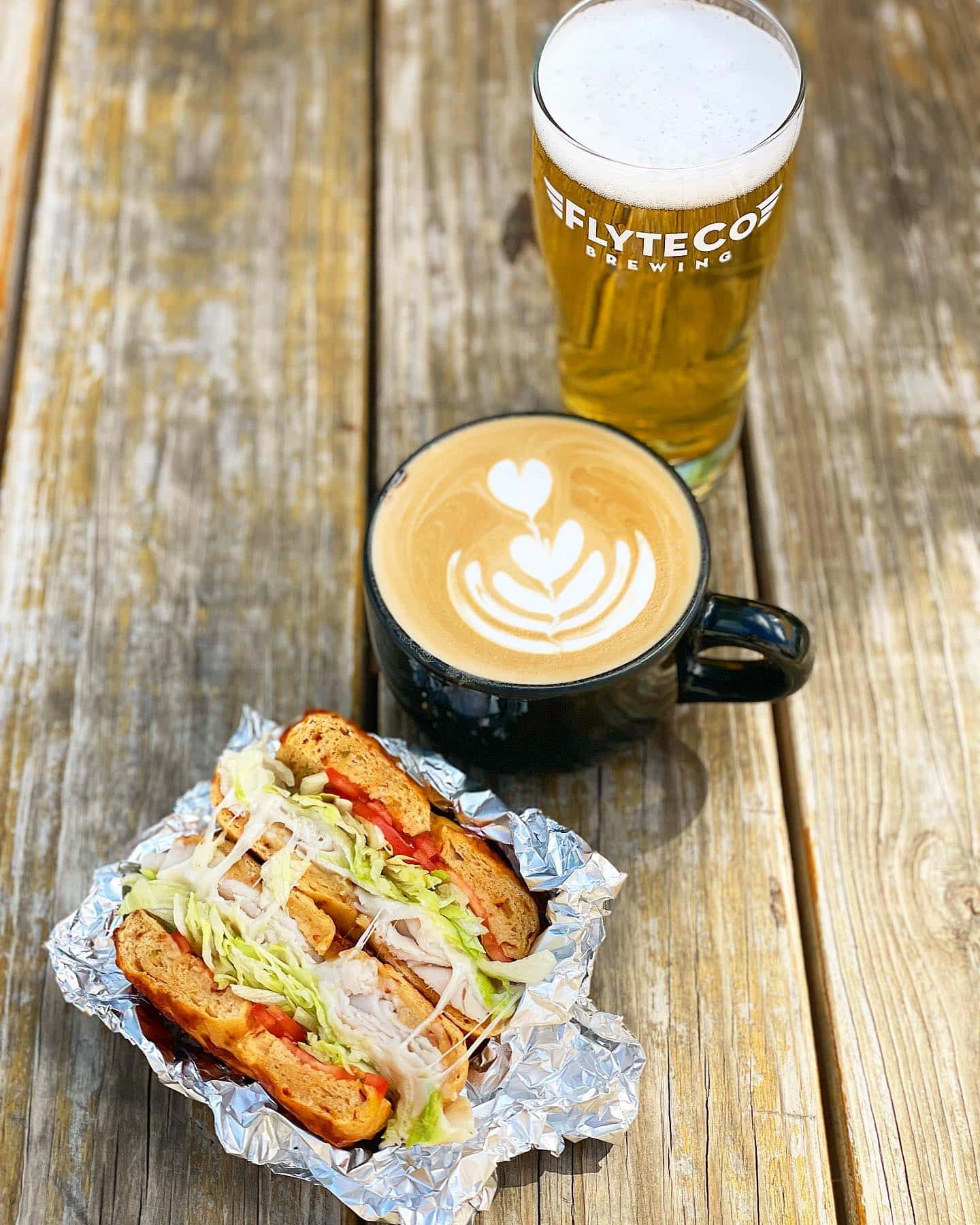 Breakfast sandwich, latte, and glass of beer all set on a wooden table at FlyteCo Brewing