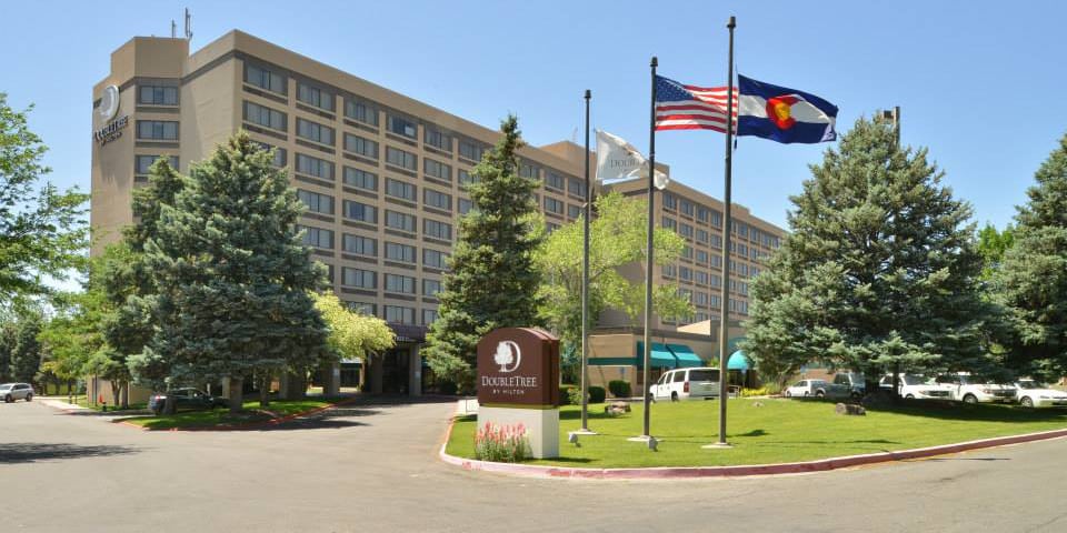 Outside of DoubleTree Grand Junction, a beige multi-story hotel building with 3 flags raised outside- a Colorado state flag, the American flag, and a DoubleTree flag