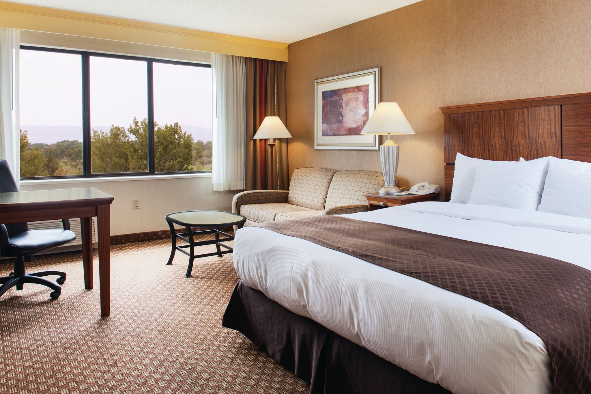 King bed in a room at the Doubletree Hilton hotel in Grand Junction. Plain king bed with white linens, a wooden dresser with a flatscreen TV, and large windows overlooking greenery