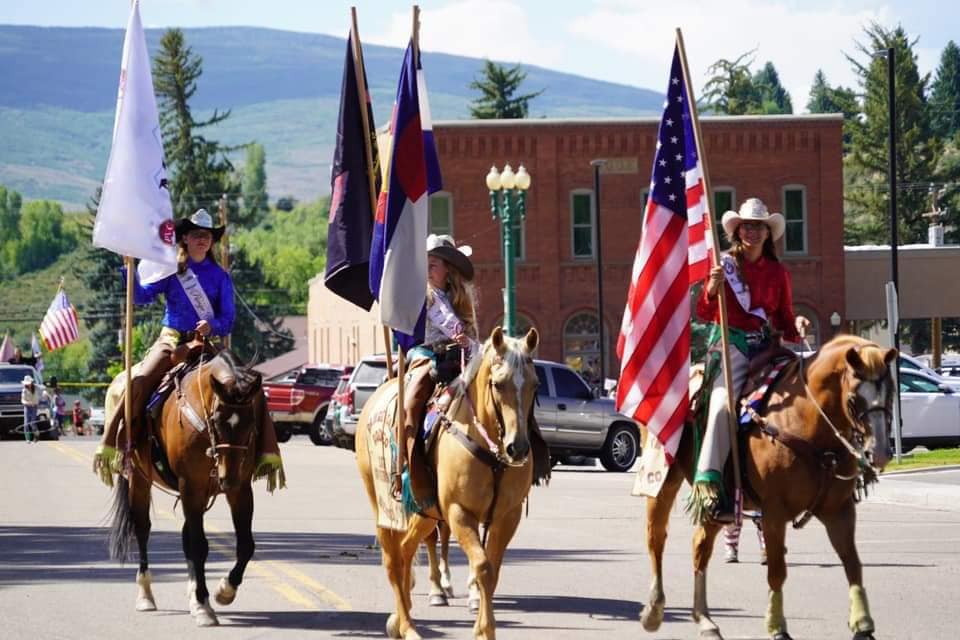 Three people riding horses down the main street of Meeker holding up American and Colorado flags for Range Call