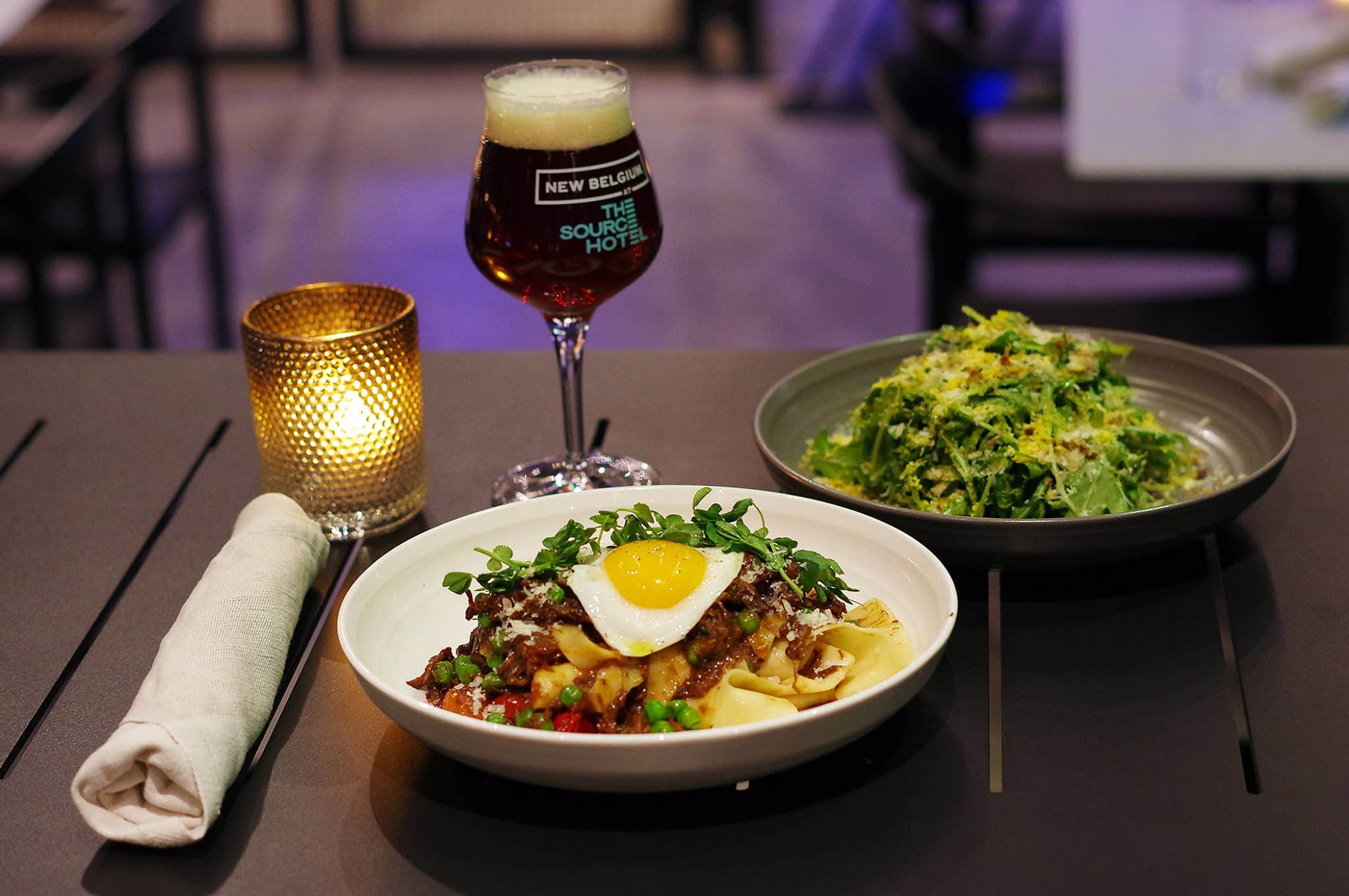 Plate of dinner food with a side salad and New Belgium Brewing beer and a candle lit. Set on a table at The Woods Restaurant within The Source Hotel in Denver