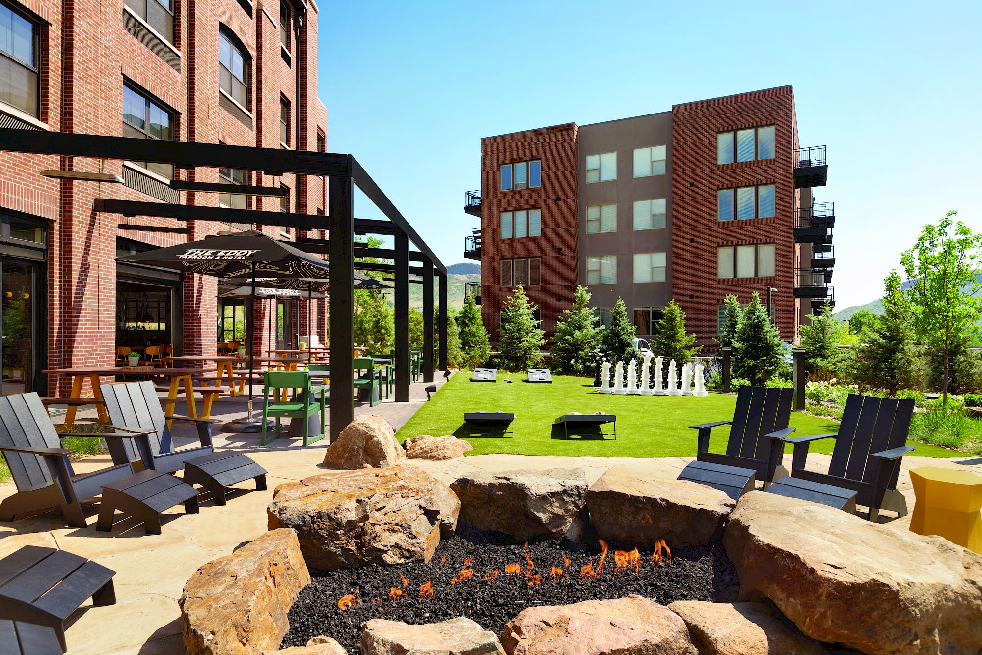 Outdoor sitting area at the Eddy Taproom & Hotel. Foreground of picture is a fire pit with black Adirondak chairs around it. Background is green turf with cornhole boards 