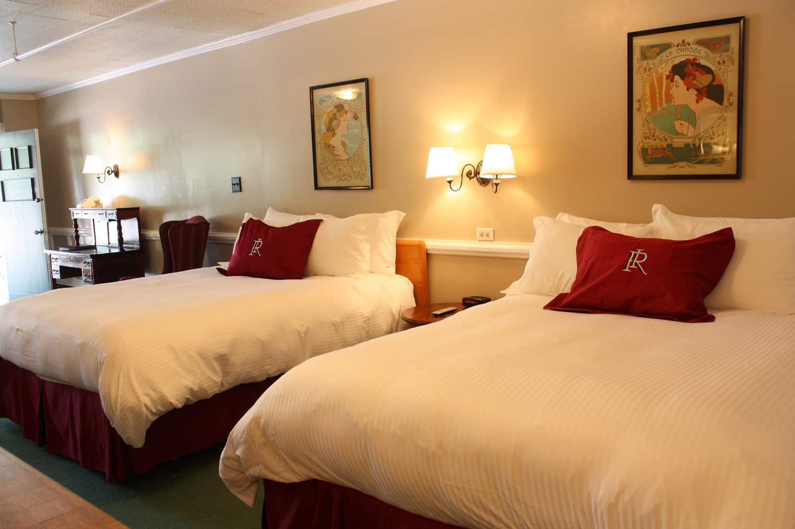 Hotel room at the Redstone Inn with two queen beds, each with white comforters and red accent pillows. Two framed pictures above each bed and a lit lamp in the middle