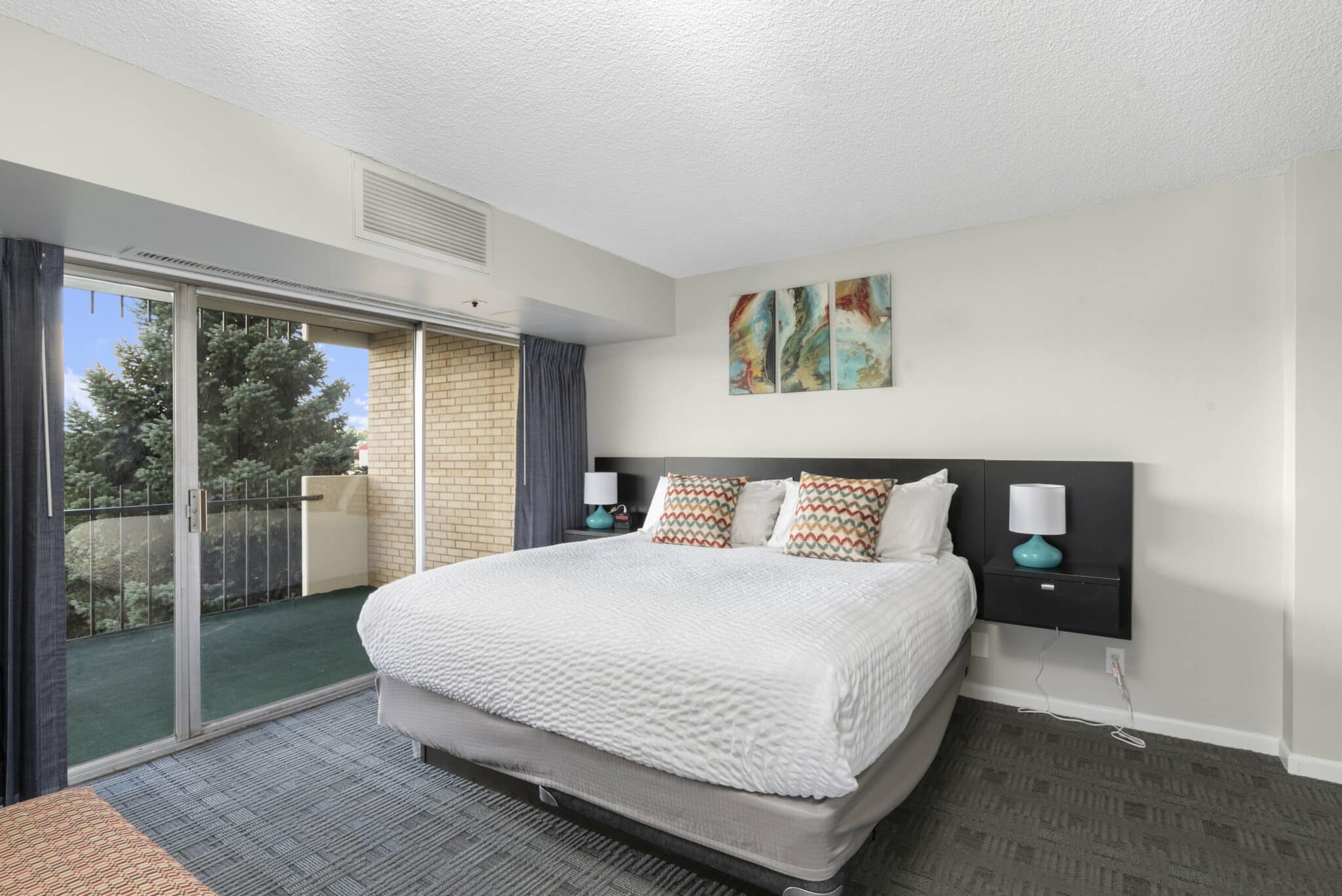 Hotel room with white linen on the bed, painted picture hung above a blue headboard, and a private balcony to the left with sliding glass doors