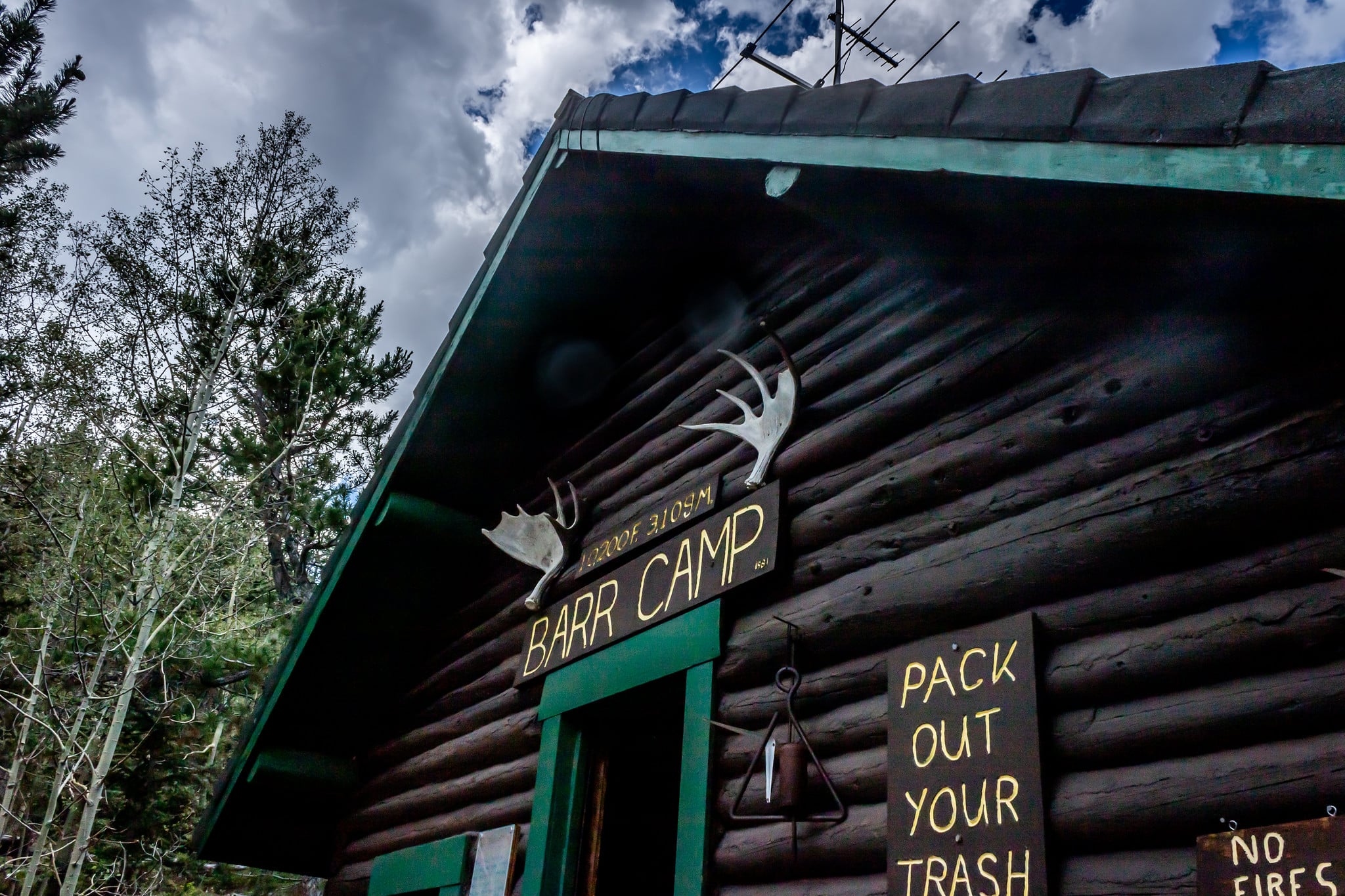 Front of Barr Camp, a dark wood log cabin with green roofing and a green door. Wooden sign with Barr Camo painted in yellow and another small sign with 'Pack out your trash'