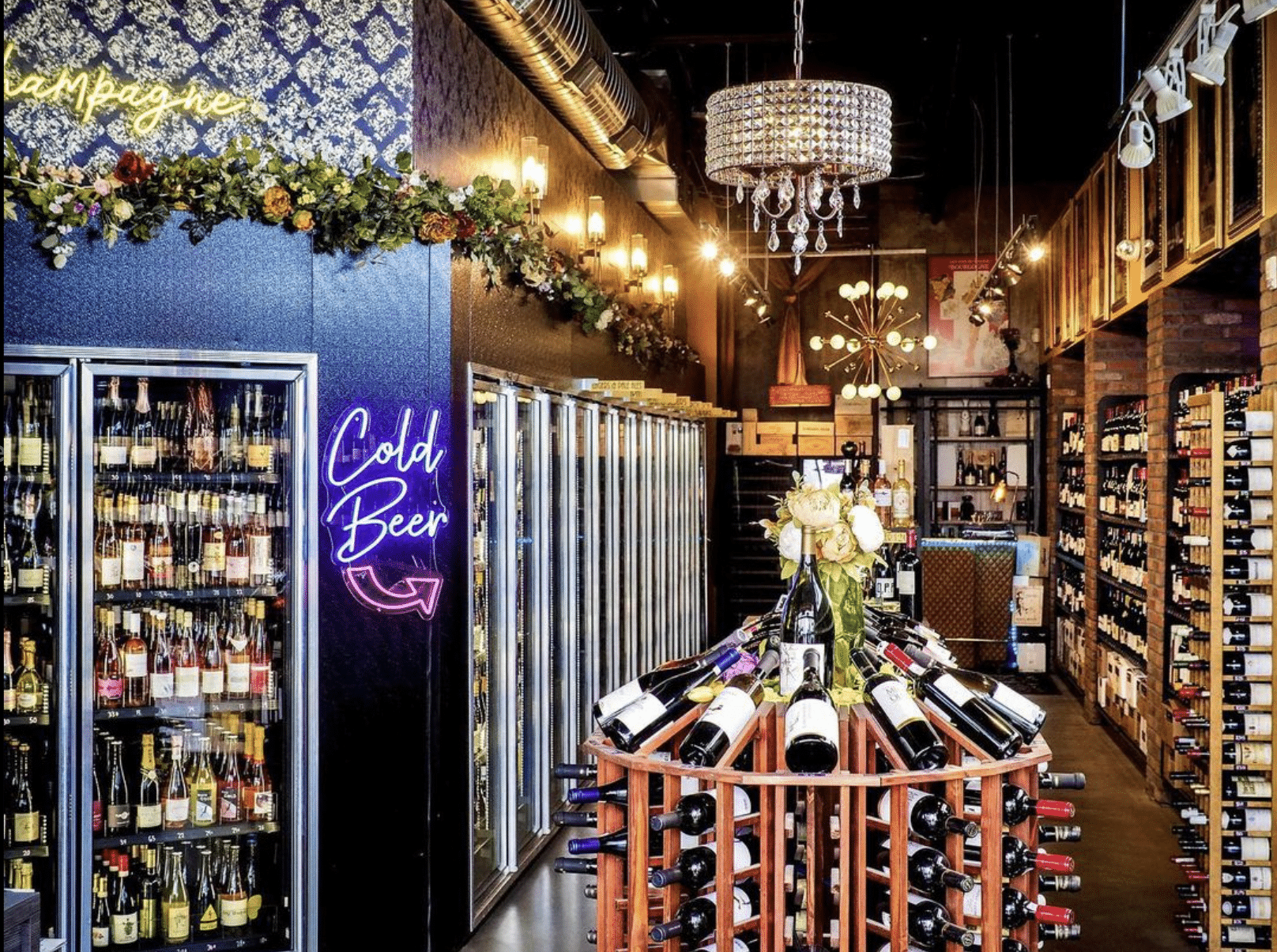 Interior of Denver Wine Merchant's store. Black walls with greenery and warm lighting throughout. Circular wine rack in the middle of the photo and a wall full of chilled beverages with a "Cold Beer" sign on the left