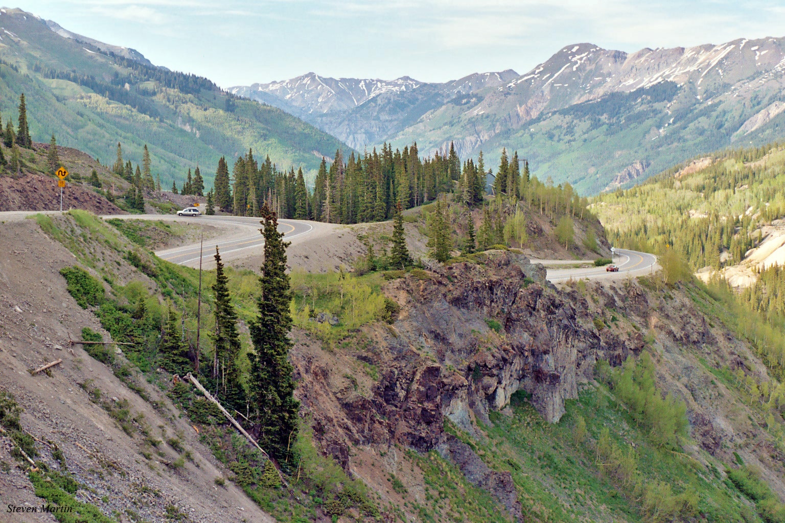 Winding road through hilly area surrounded by large mountains and trees. Pictured is the Million Dollar Highway section of the San Juan Skyway National Scenic Byway