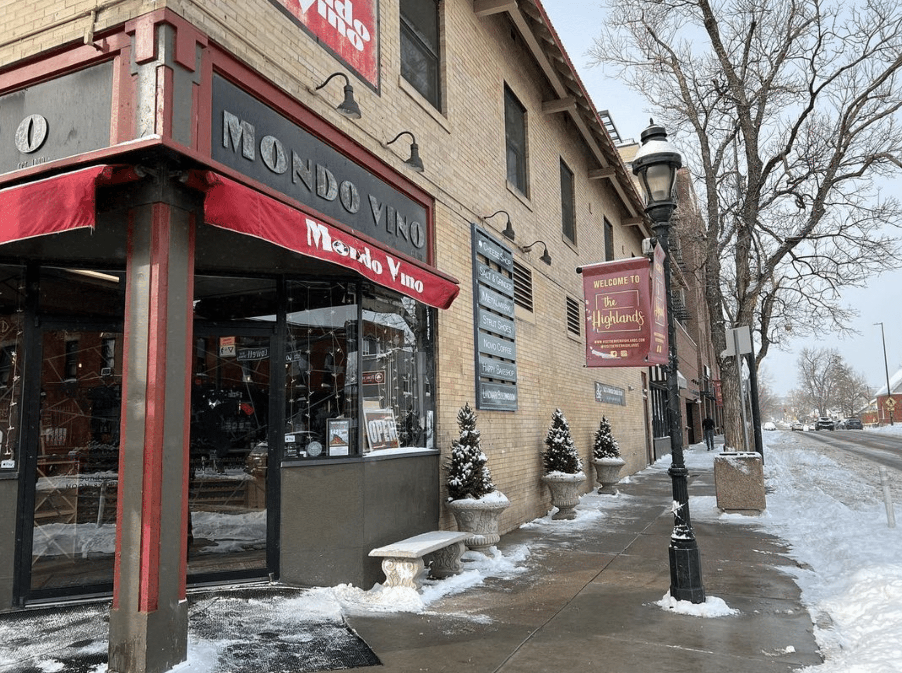 Corner storefront for Mondo Vino in Denver. Red cloth awning with the shop name written in white letters and a light dusting of snow on the sidewalk in front