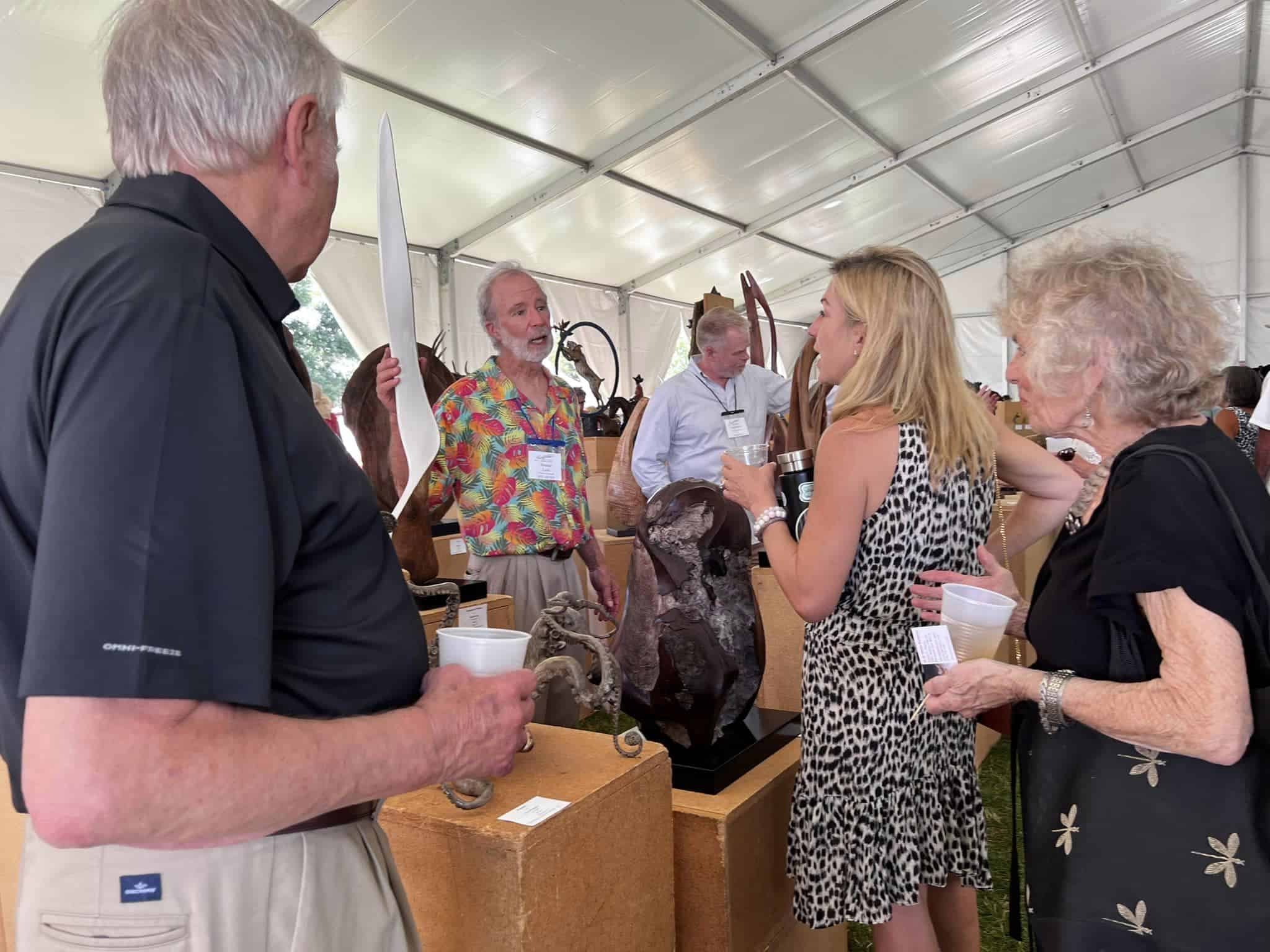 Multiple people under a large white tent, mingling with artists at the Sculpture in the Park Patron Party.