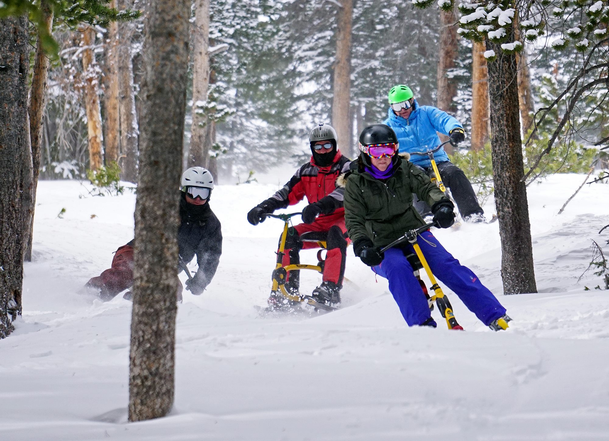 4 people in snow gear on bright yellow snowbikes going down a snowy ski slope in Breckenridge.