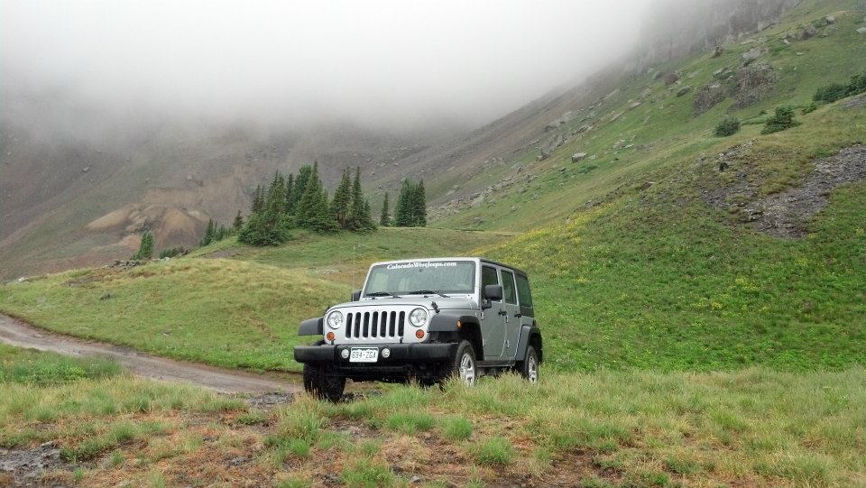 Silver Jeep parked in the middle of a foggy alpine grass meadow in a valley.