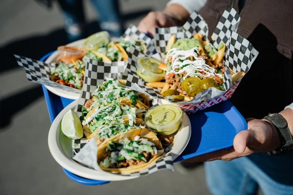 person carries 3 plates of tacos on a blue food tray at the Aurora Borealis Festival in Colorado