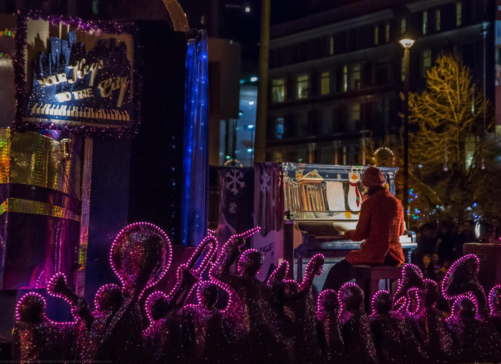 woman in orange winter jacket playing piano at night surrounded by Christmas Lights in Downtown Denver