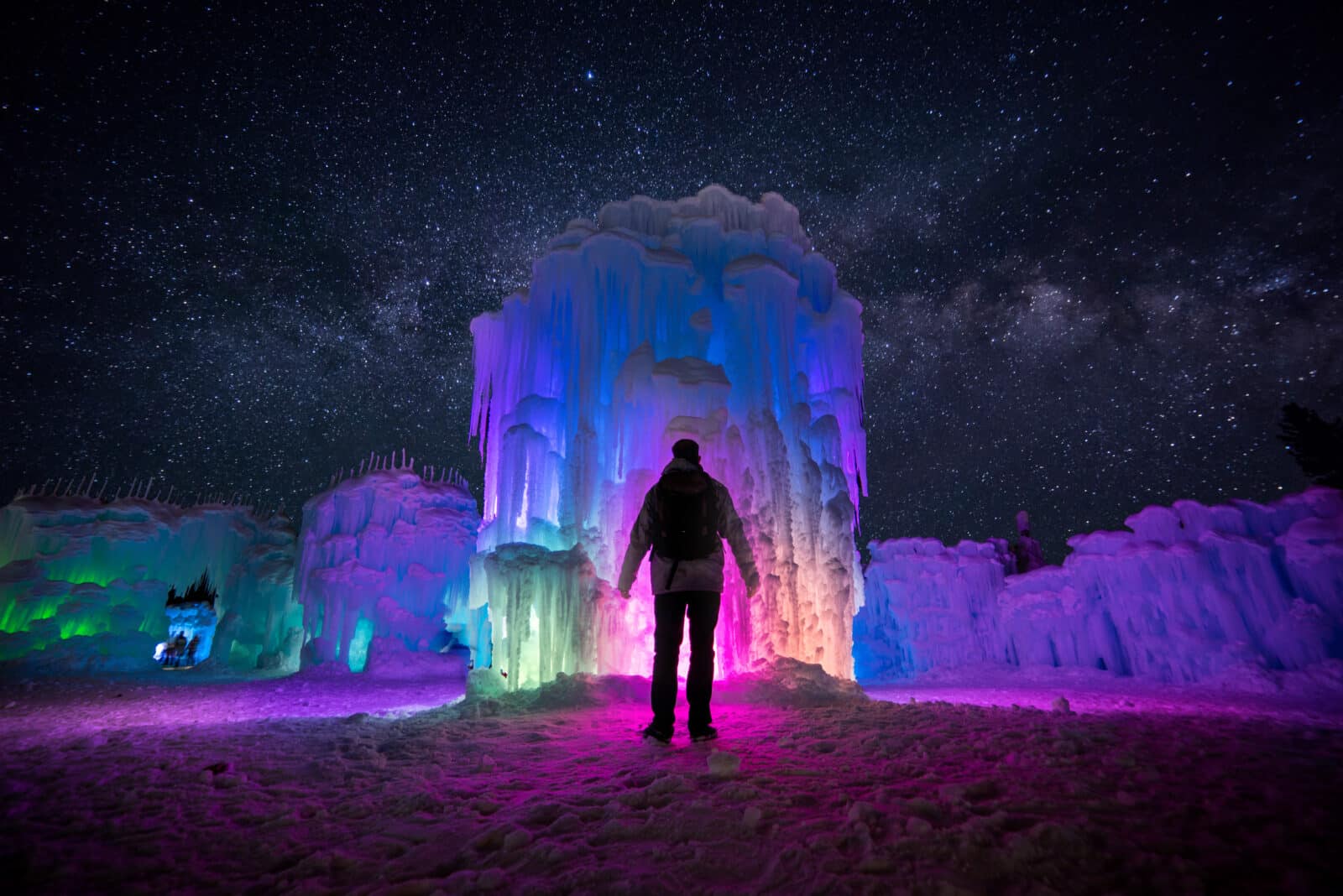 man stands in front of a giant ice castle that is lit up with many colors at night